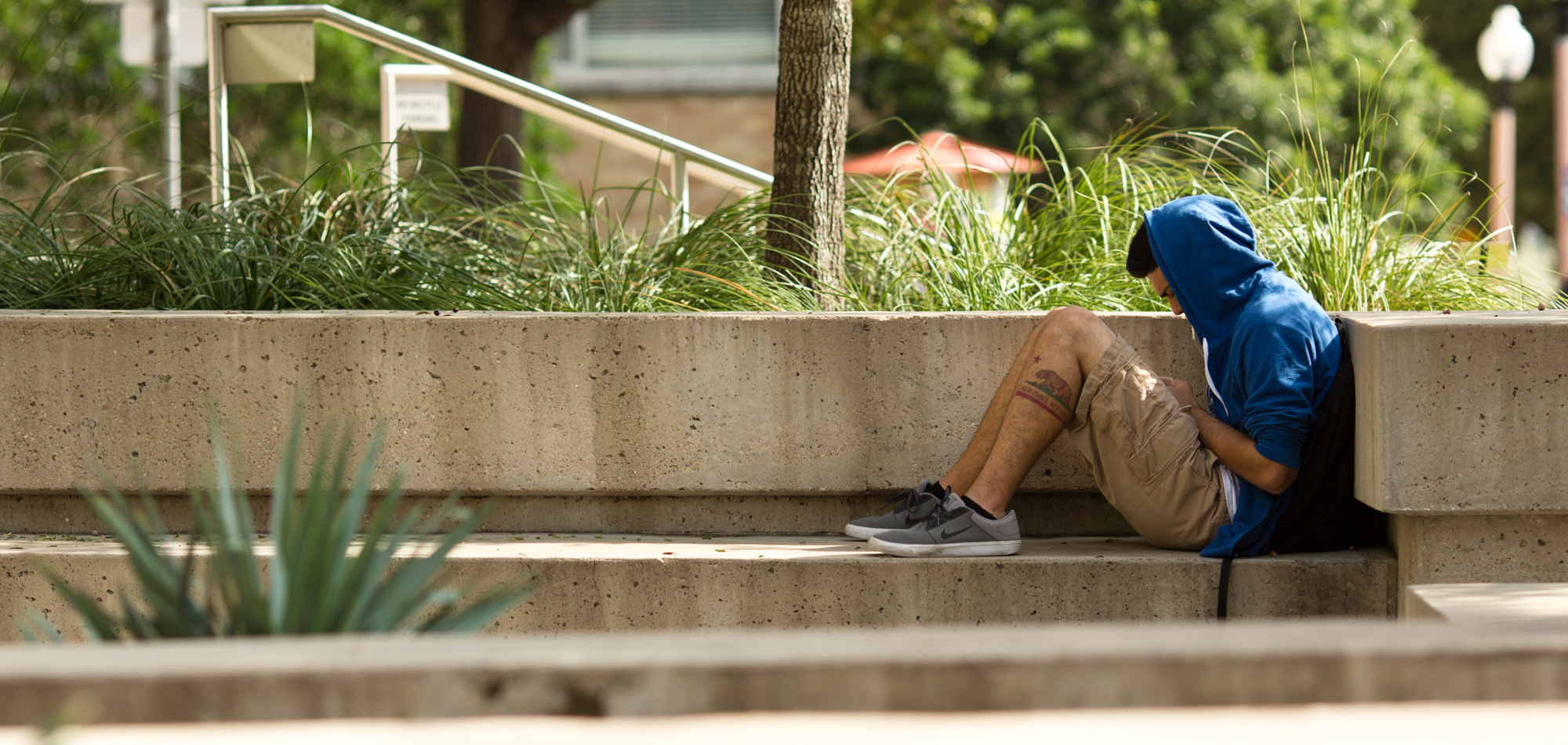 Young man in a blue sweatshirt and shorts huddles on a long concrete bench