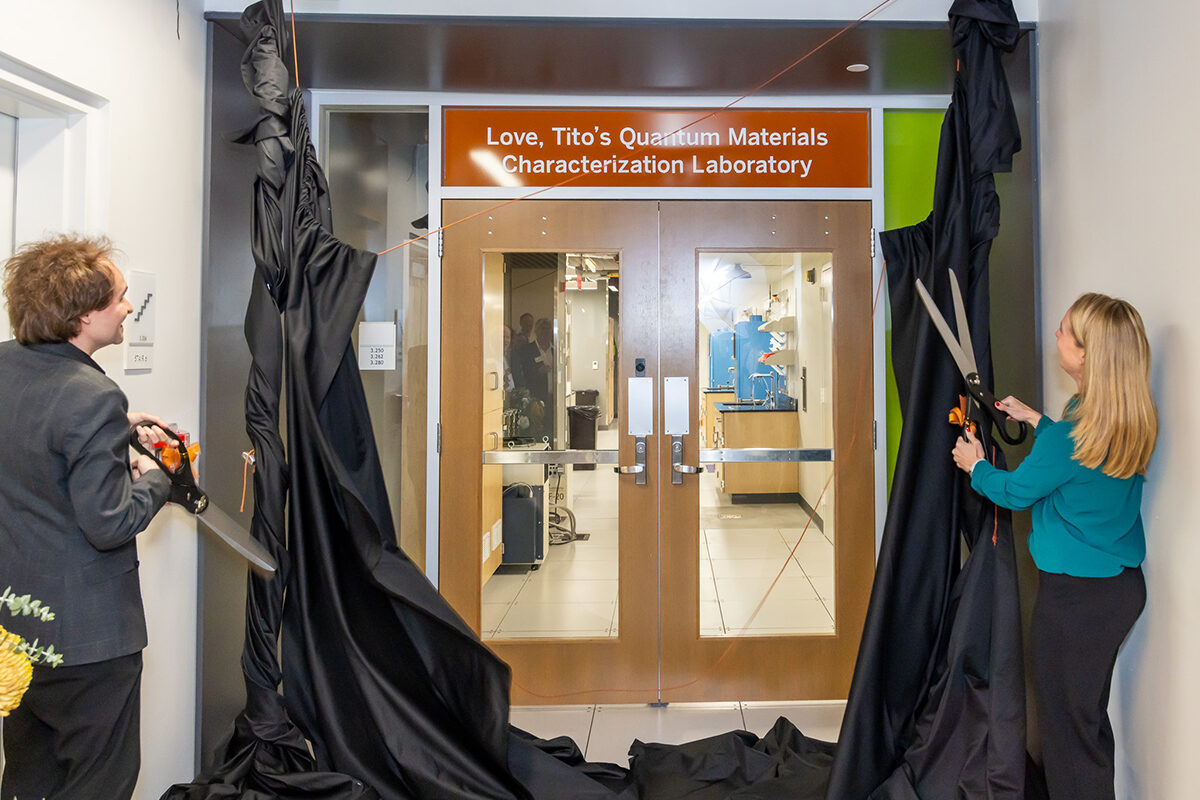 Two people with large scissors cut the cords on a pair of black drapes, revealing a new sign over the entrance to a lab. The sign reads: " love tito’s quantum materials characterization laboratory."